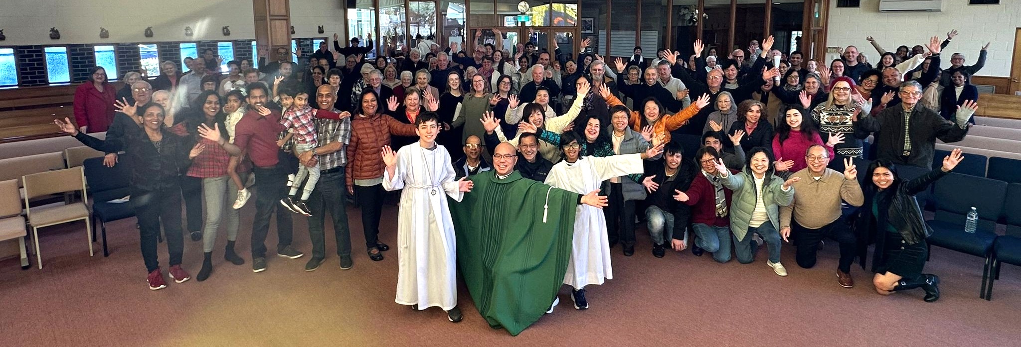 The parish community of St Timothy's gathered after Mass, hands raised in welcome, with Father Alex Chow and two altar servers at the centre